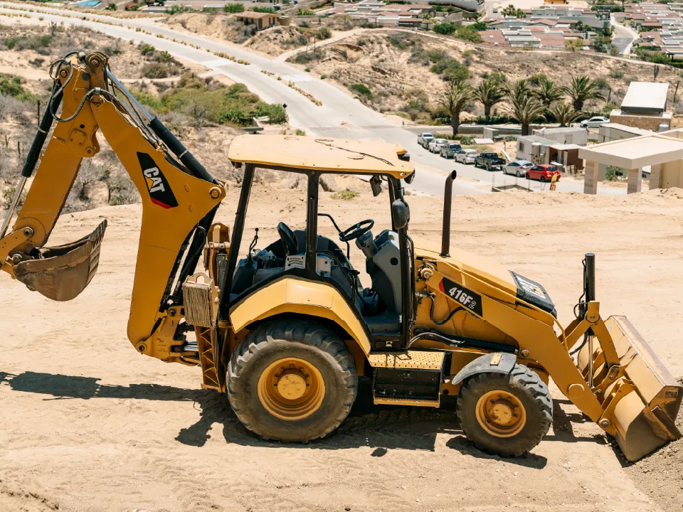 CAT 416F2 backhoe loader on a graded hillside lot, looking down at a street lined with parked cars