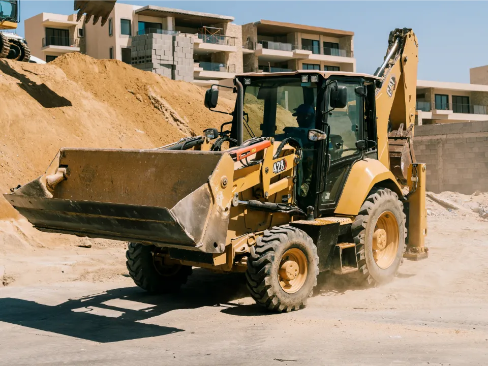 CAT 428 backhoe loader driving past a sand pile, its operator at the controls, with new apartment buildings behind
