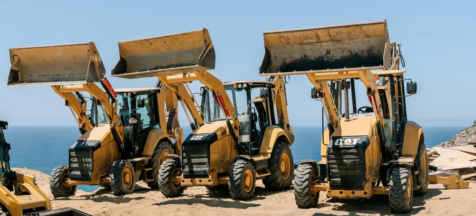Three yellow CAT backhoe loaders lined up on a sandy bluff with their front buckets raised and the ocean behind them