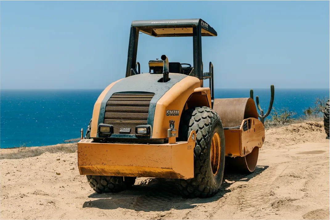 Front view of the CASE roller parked on sand at the edge of a cliff, showing its engine grille, lights, chunky rear tires and drum, with the deep blue sea and a tall cactus behind