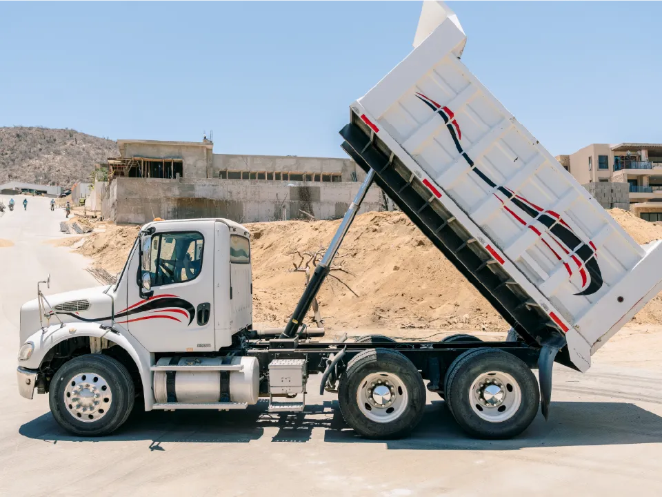 White Freightliner dump truck with its bed raised on a paved street beside a building site