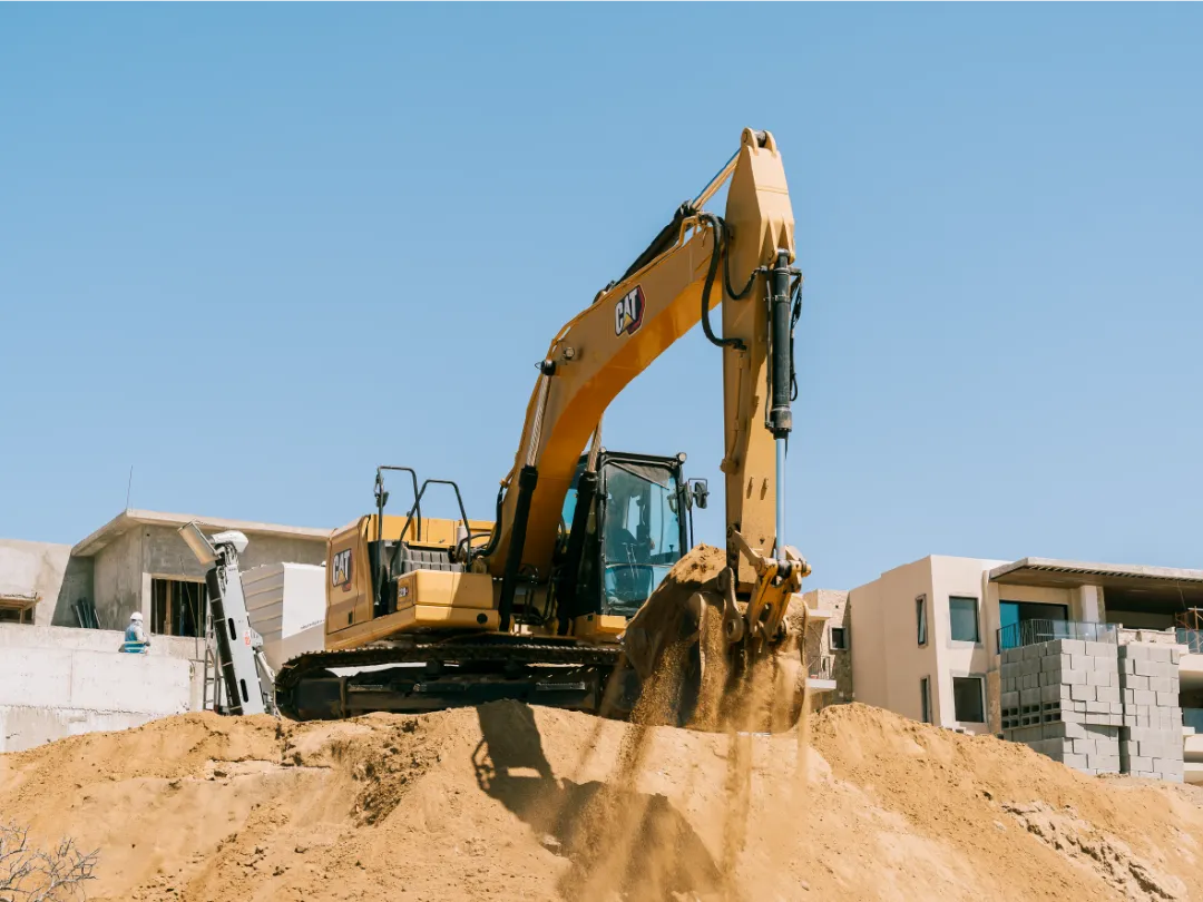 CAT 320GC excavator facing the camera, sand pouring from its raised bucket in front of the cab, with a block apartment building in the background