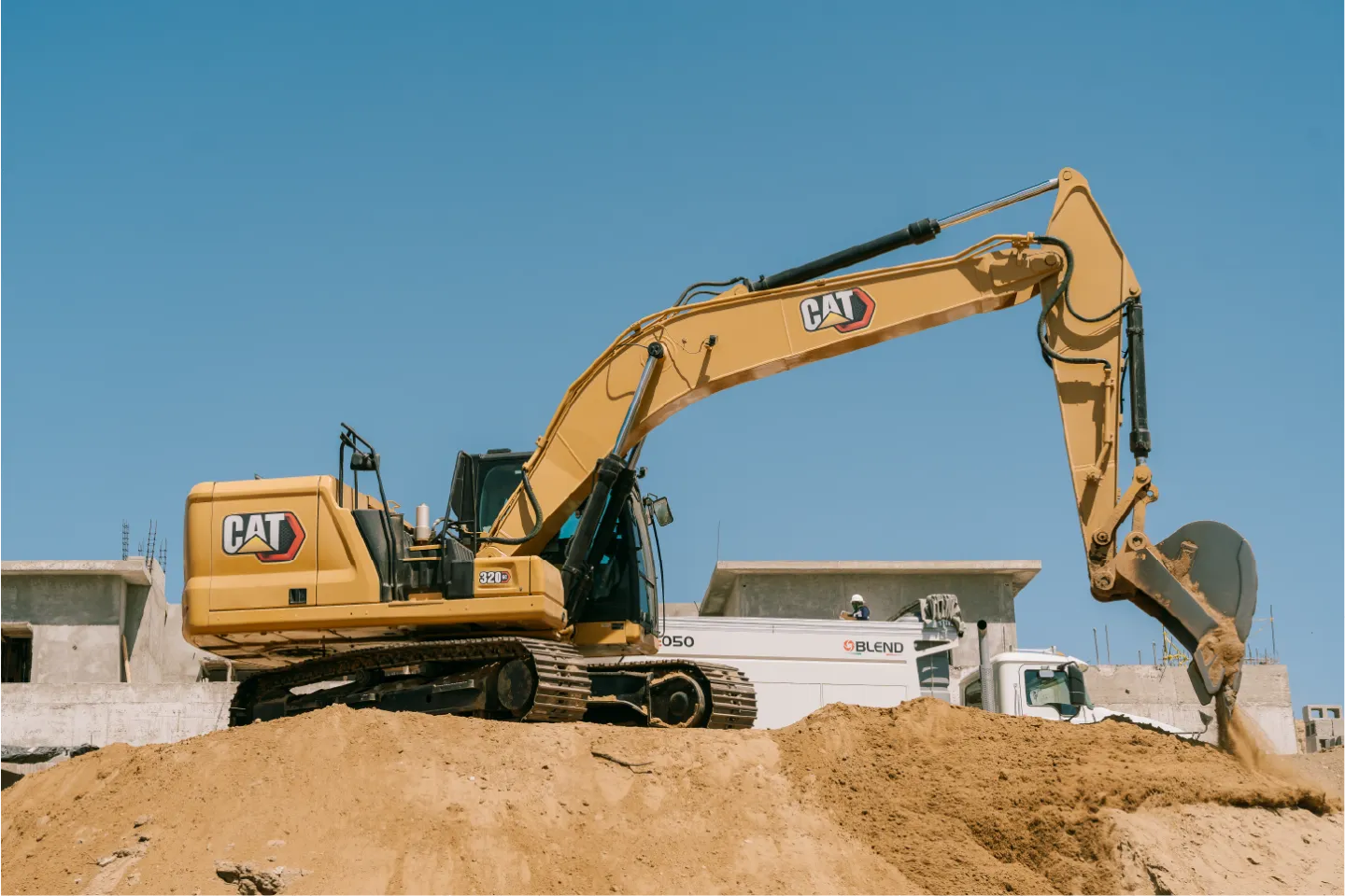 Yellow CAT 320GC crawler excavator on a sand mound, arm stretched to the right and bucket lowered, with a white work truck and unfinished houses behind it