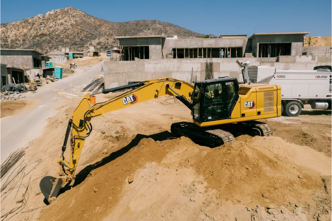 CAT 320GC excavator perched on a dirt mound, reaching its arm down the slope toward a street, with concrete homes and a rocky hill behind