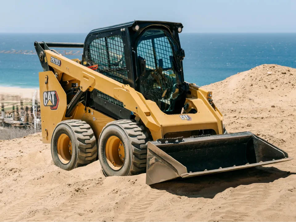 CAT 262D3 skid steer loader with a wide bucket, sitting in loose sand on a bluff above the beach