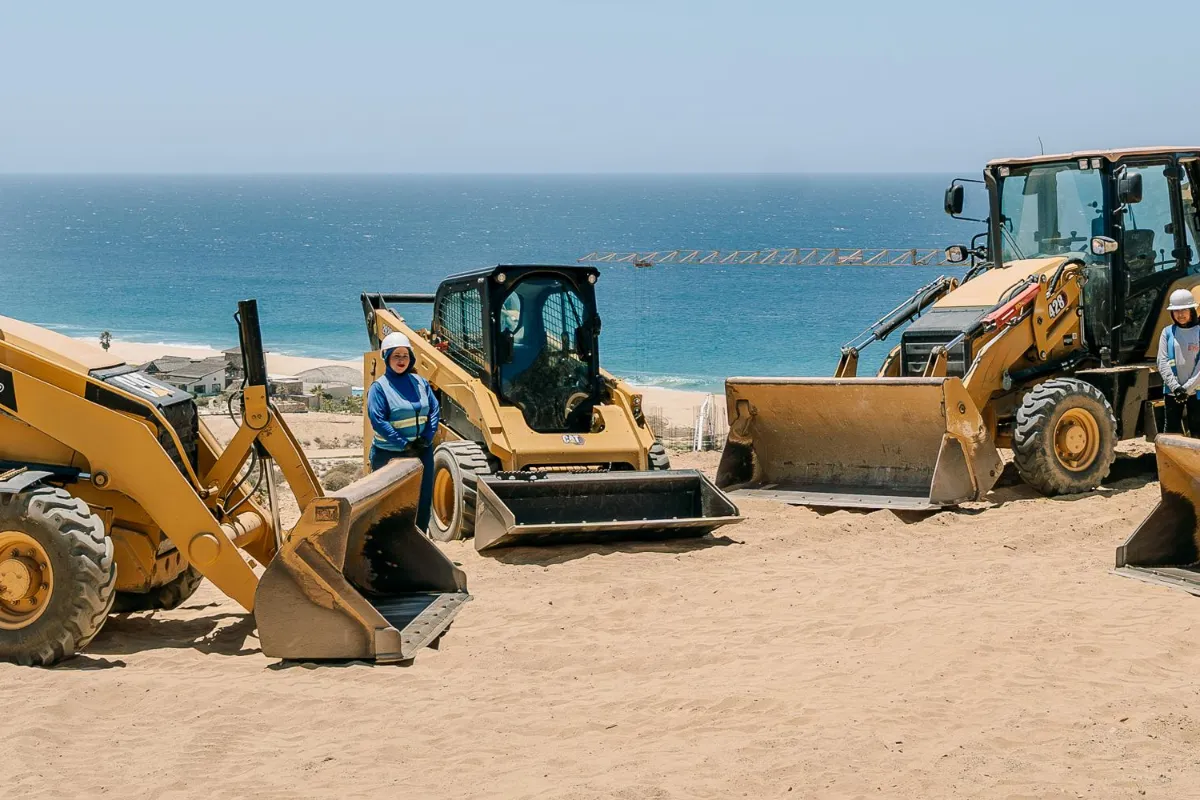 Creatio crew member in a hard hat and safety vest standing beside the compact CAT skid steer, parked between two larger CAT backhoe loaders on a sandy lot by the sea