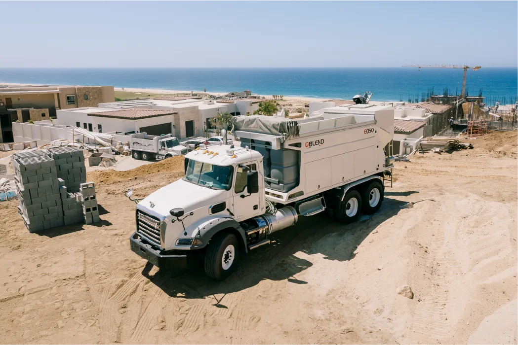 Blend E050 mixer truck on a sloping residential building site beside stacks of concrete block, with new houses and the ocean behind