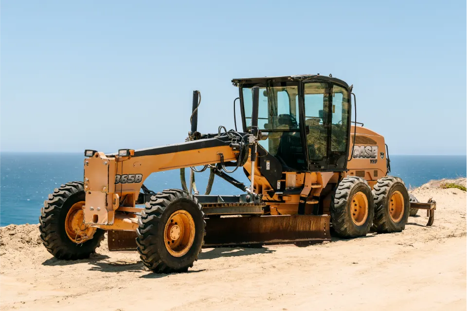 Angled view of the CASE grader showing its moldboard and rear ripper, standing on sand against the ocean