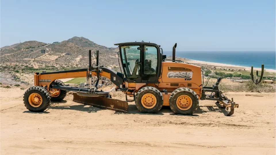 Side view of the CASE 865B grader on a cleared dirt lot, with desert hills, a cardon cactus and a long beach behind it