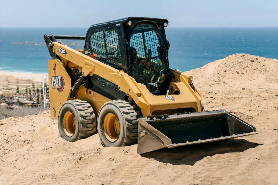 Angled view of the CAT skid steer, rubber tires and bucket down on a sand ridge, a beach and resort buildings behind