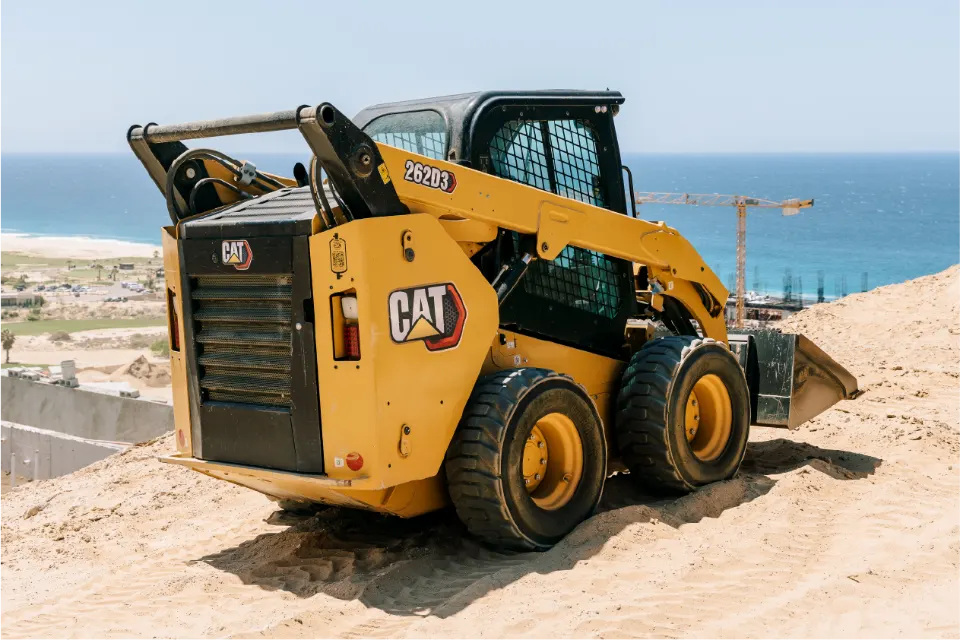 The CAT 262D3 from behind, rubber tired and yellow, at the top of a sand slope with a tower crane and the sea in the distance