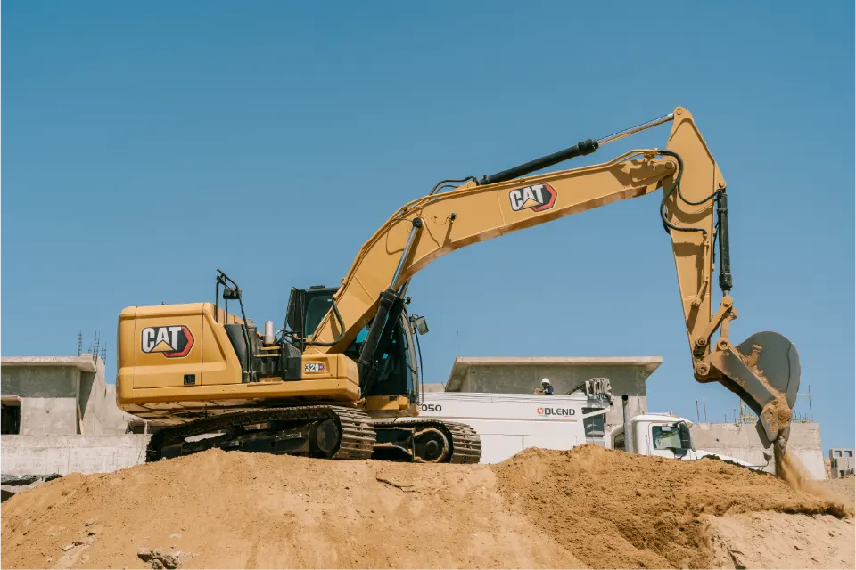 CAT 320GC excavator extending its arm across a mound of sand, with a Blend mixer truck and concrete buildings in the background