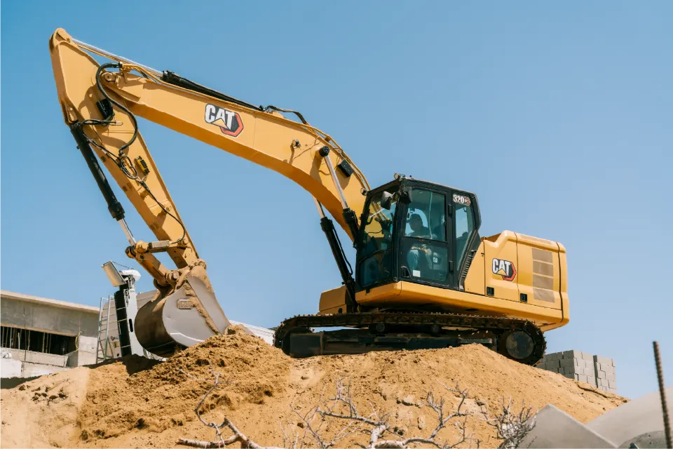 Low angle view of a CAT 320GC excavator holding its bucket high over a sand mound, dry branches in the foreground