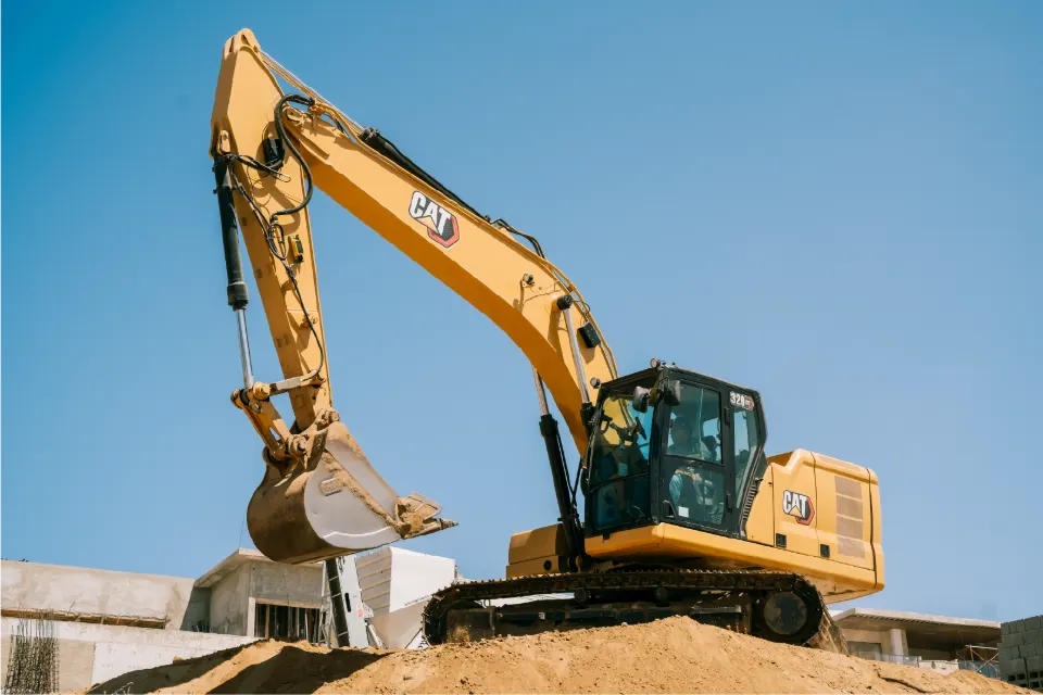 Low angle view of a CAT 320GC excavator atop a sand mound, arm curled and bucket lifted, concrete buildings at the edge of the frame