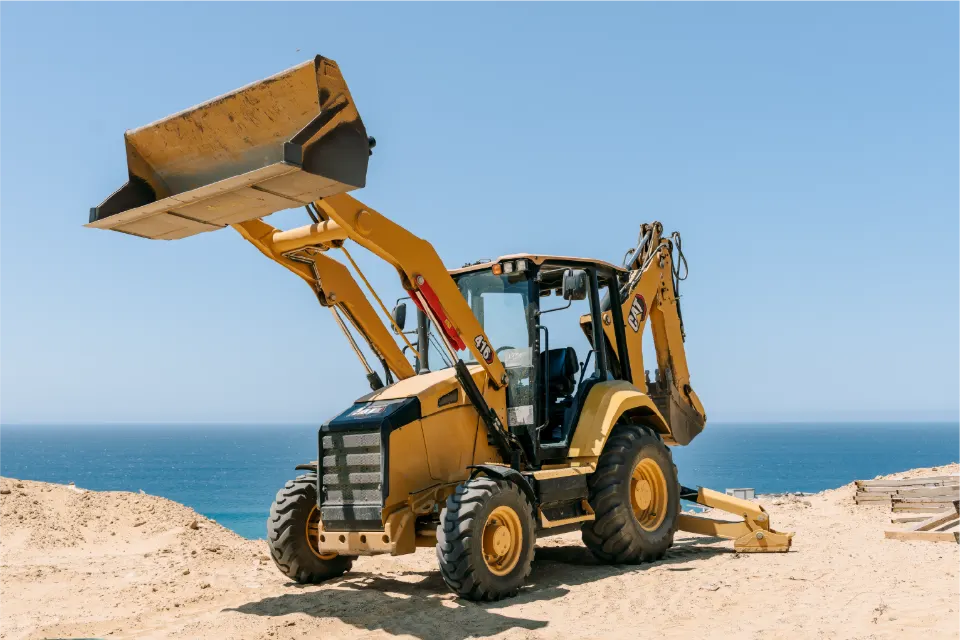 A CAT 416 backhoe loader holds its loader bucket above the cab on a sandy lot, the sea behind