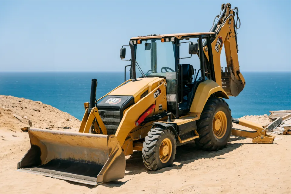 CAT 416 backhoe loader, front bucket resting on the ground, right where a bluff drops toward a blue sea