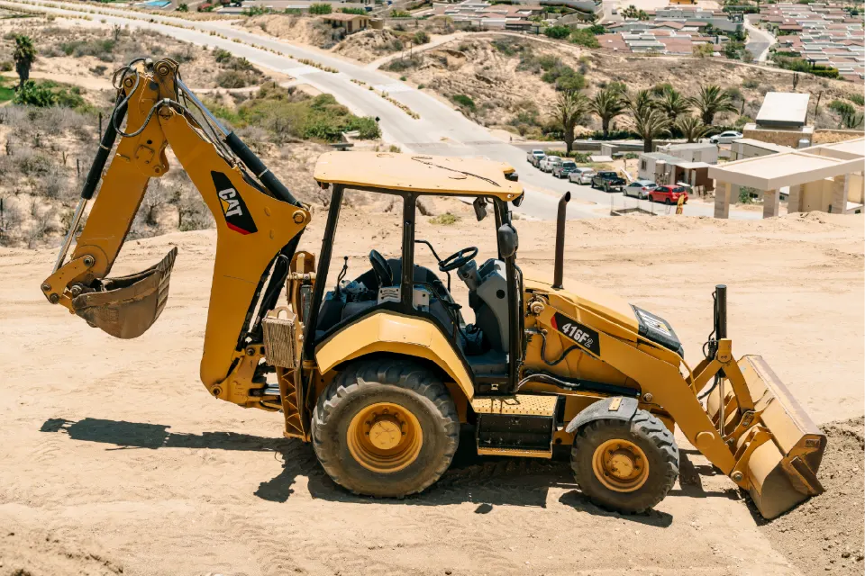 Sloping lot above a neighborhood with a CAT 416F2 backhoe loader at work, a winding road and parked cars in the distance