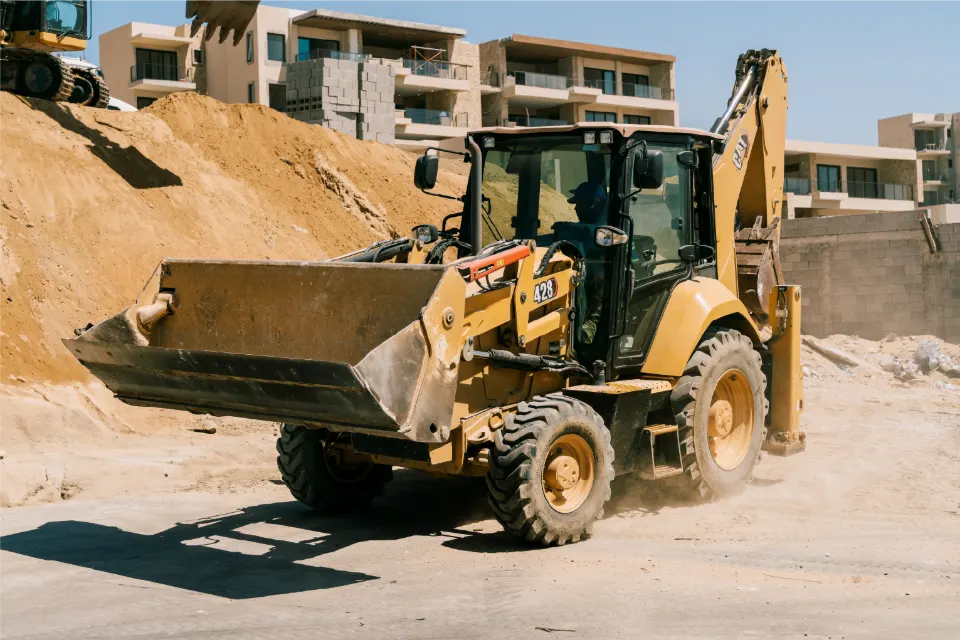 Operator aboard, a CAT 428 backhoe loader rolls across a site with its front bucket lifted, below a sand cut and apartment buildings