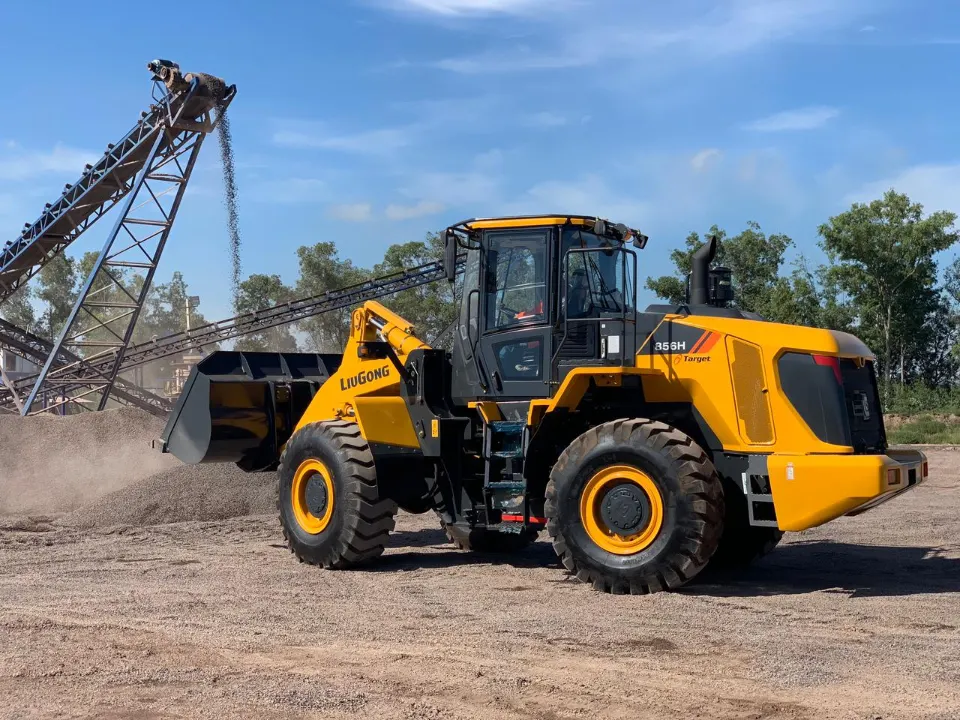 LiuGong 856H wheel loader, bucket lowered close to the ground, beside a conveyor tower with leafy trees and blue sky behind