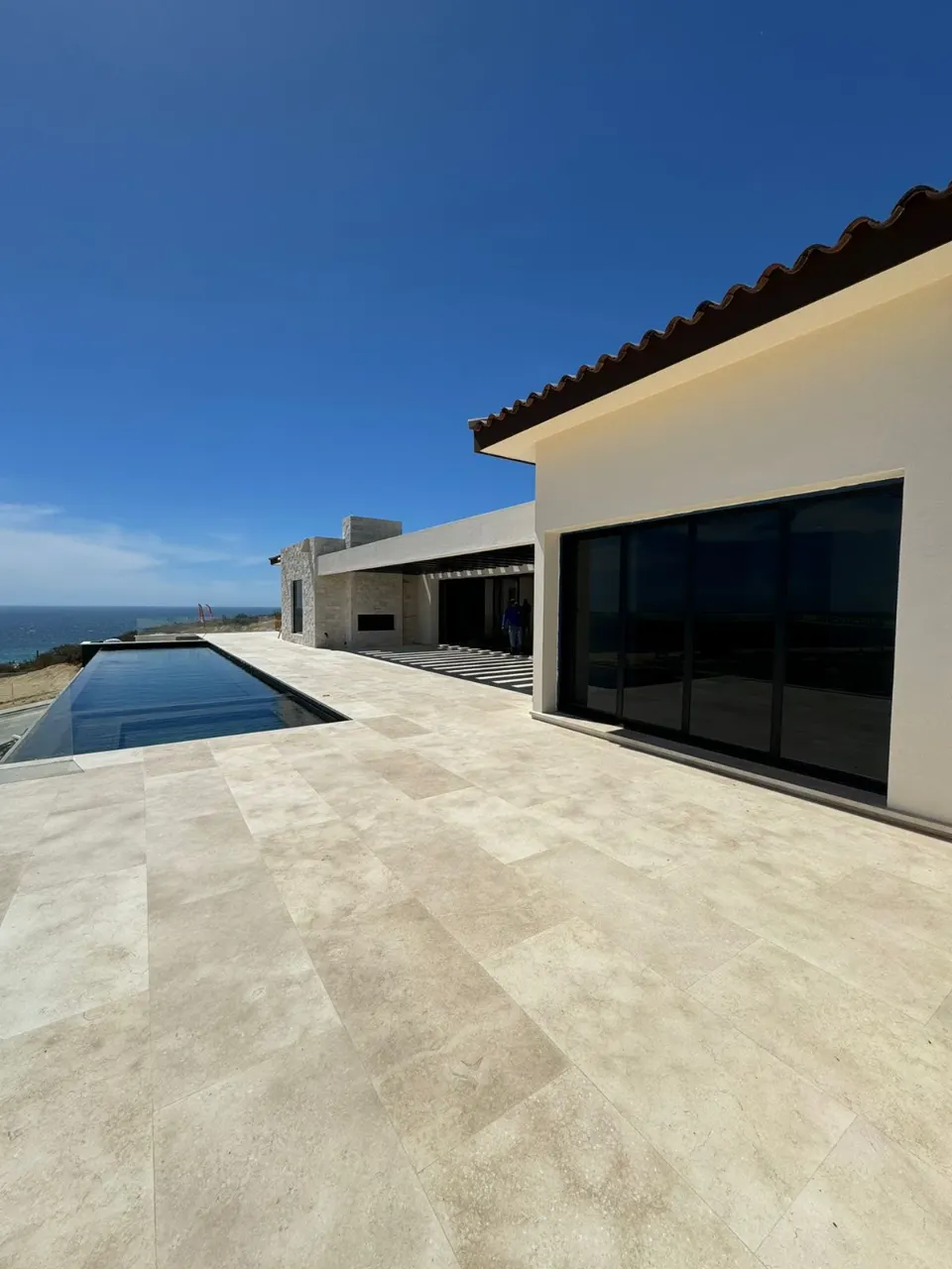 Bright white house with dark window walls, a clay tile roof edge and a long pool beside a travertine deck under a blue sky
