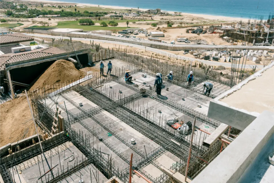 Workers tying a grid of reinforcing steel inside a large excavated foundation, with a sand cut on one side and a golf course and coastline in the distance
