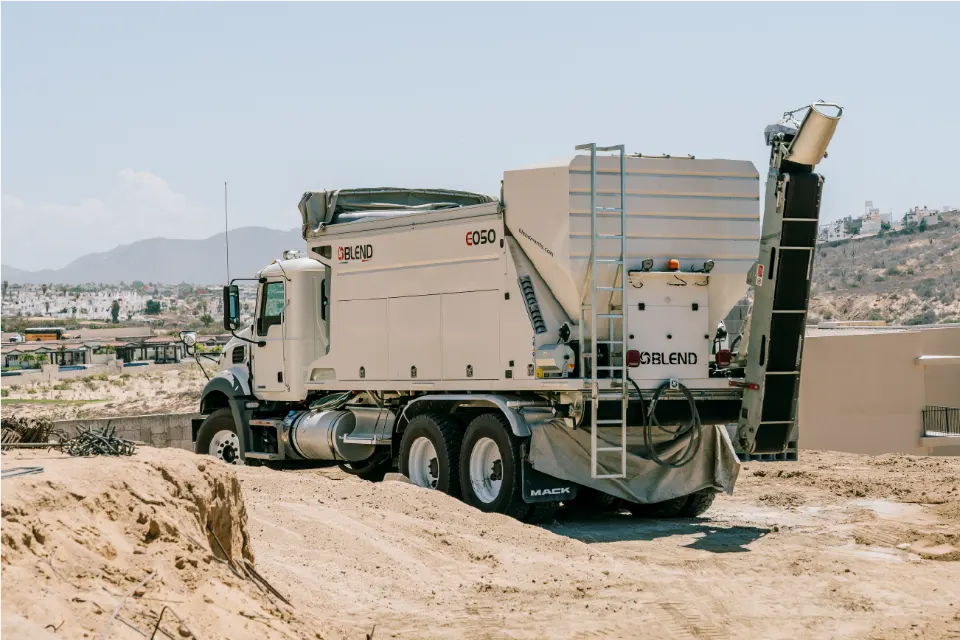 The Blend E050 mixer on a Mack truck, seen from behind on a sand mound, conveyor raised, a town and hills beyond