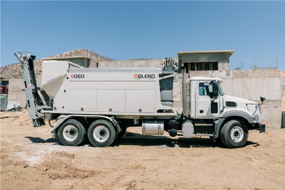 Side view of the white Mack truck and Blend E050 mixer with its rear conveyor raised, parked in front of concrete walls on a residential build