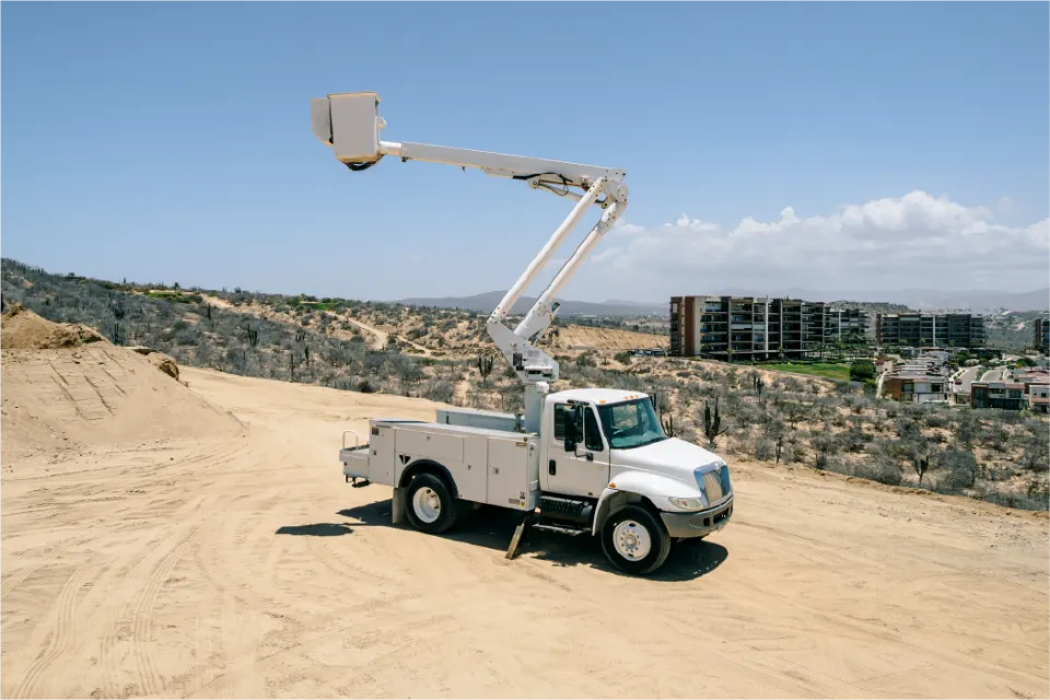 Basket lift on an International 4300 truck, parked on a sloping dirt lot with the boom up, coastal scrub and a row of apartment buildings behind