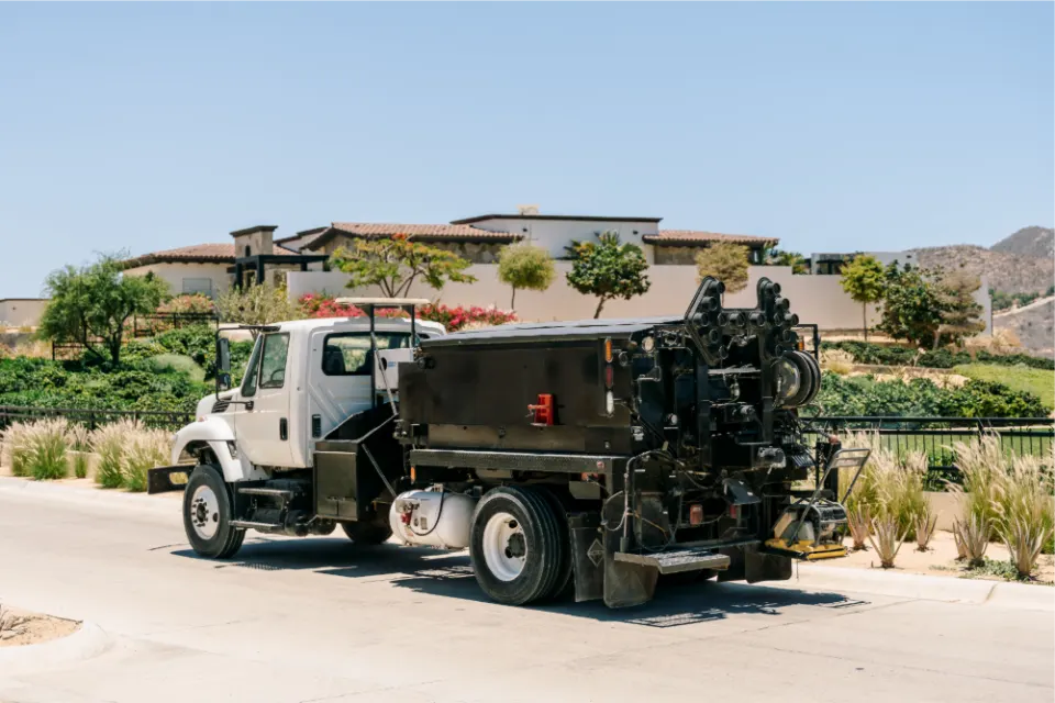 Pothole patcher truck parked on a curving residential street lined with ornamental grasses, with tile roofed villas and hills behind