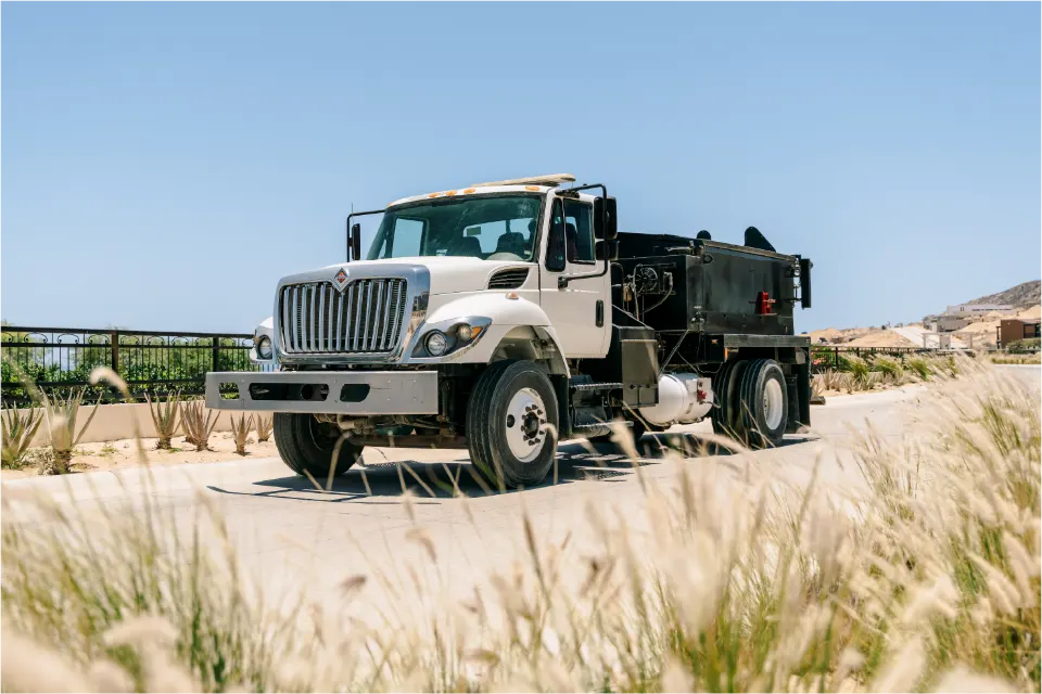 Angled front view of the white International patcher truck parked on a ridge road, tall dry grass in the foreground and a railing behind