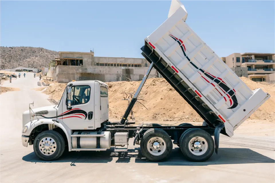 Profile of a white Freightliner M2 dump truck with its dump body raised high, new buildings and sandy slopes around it