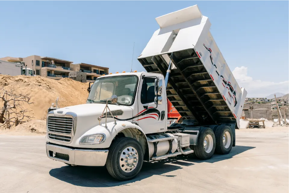 Angled front view of a white Freightliner M2 dump truck, body lifted, with sand banks and new buildings behind