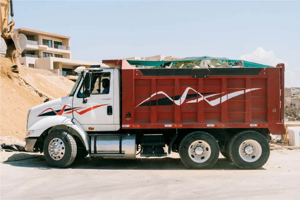 White International TranStar dump truck, its red dump body full of debris under a green tarp, a backhoe arm and apartment blocks behind