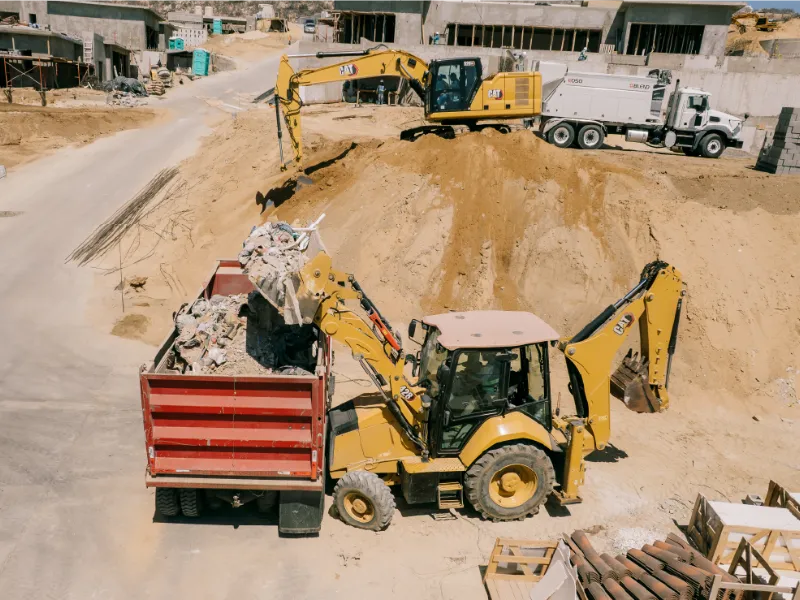 Retroexcavadora CAT cargando escombro en un camión de volteo rojo en una obra residencial
